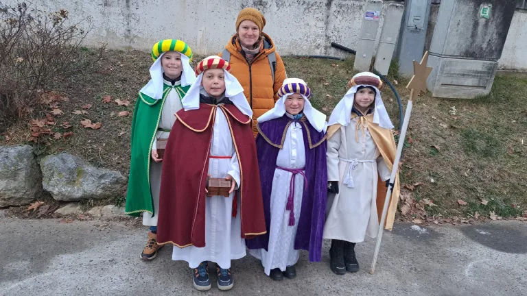 Fünf Kinder in verschiedenen Kostümen stehen in einer Reihe auf einer Straße, eines hält eine Schachtel, und eine Frau mit Mütze steht hinter ihnen.