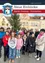 Eine Gruppe von Menschen, darunter Erwachsene und Kinder, posiert für ein Foto vor einem Gebäude mit einem Weihnachtsbaum und einem Uhrenturm. Das Bild hat einen Wasserzeichen oben.