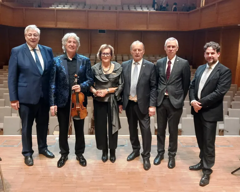 Five people are standing on a stage in a theater. A man in the middle is holding a violin. The people are wearing formal attire. Behind them are rows of chairs.