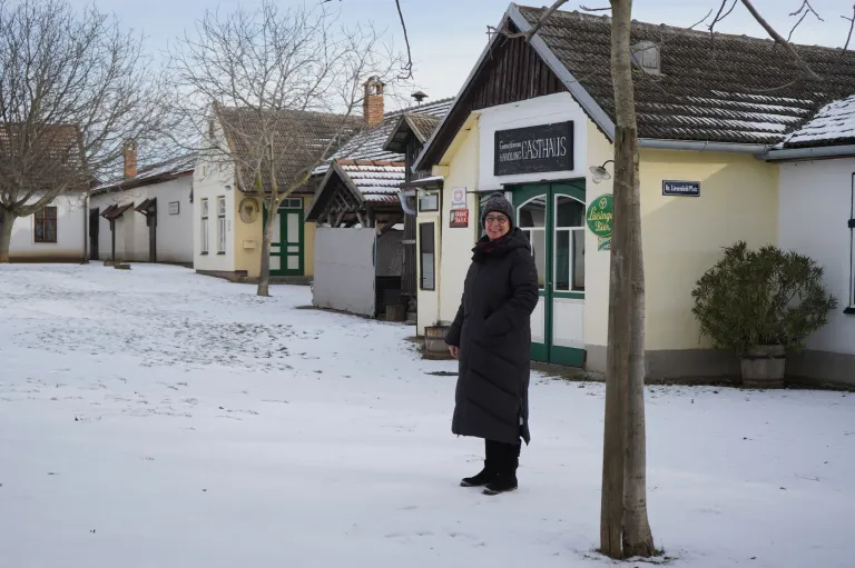 A woman in a black coat and boots stands in the snow in front of a small building with a sign that reads 'Casthaus'.