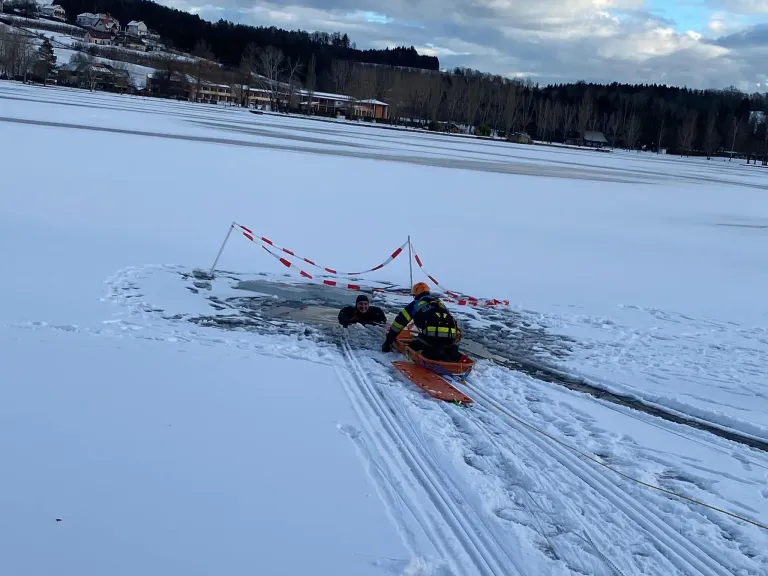 Zwei Personen in Winterkleidung auf einem verschneiten See. Eine klettert aus dem Wasser, während die andere auf einem Schlitten sitzt, beide tragen Helme.