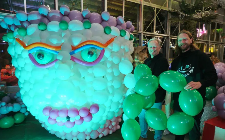 Zwei Männer stehen neben einer großen Ballonskulptur. Die Skulptur zeigt ein farbenfrohes Gesicht mit Augen, Nase und Mund aus Luftballons. Der Mann auf der rechten Seite hält eine Handvoll grüner Ballons. Dahinter ist ein verschwommenes Bild eines Dinosauriers zu sehen.