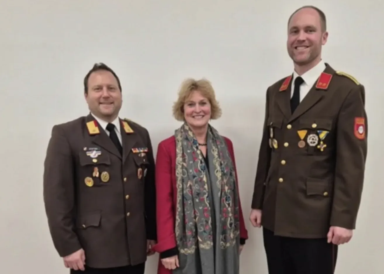 Three individuals, two men and a woman, are standing together. They are all wearing formal attire. The two men are in military uniforms with medals on their chests. The woman is wearing a red jacket and a scarf. They are all smiling and seem to be posing for a photo.