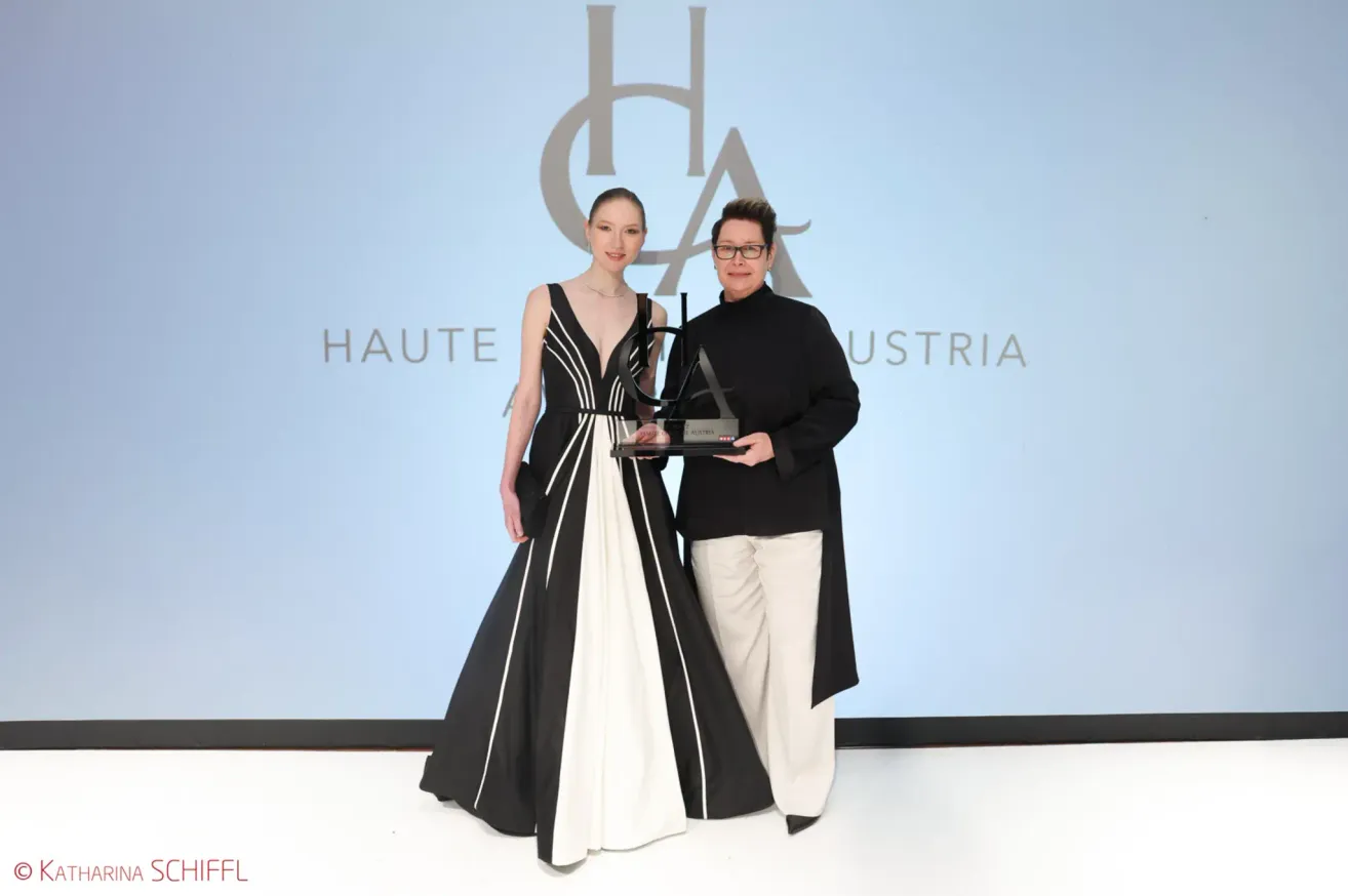 Two individuals stand on a stage holding an award, with one in a black and white dress and the other in black with glasses. Behind them is a backdrop with the text 'HAUTE AUSTRIA'.