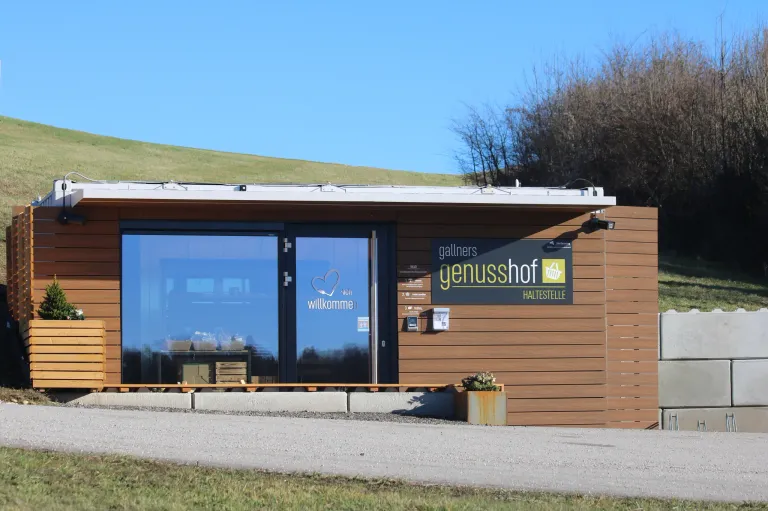 Ein modernes Gebäude mit Glastüren und einer hölzernen Außenfassade. Es trägt ein Schild mit der Aufschrift 'genusshof HALTESTELLE' und einem Herzsymbol. Vor dem Gebäude steht eine kleine Pflanze in einem Metalltopf.
