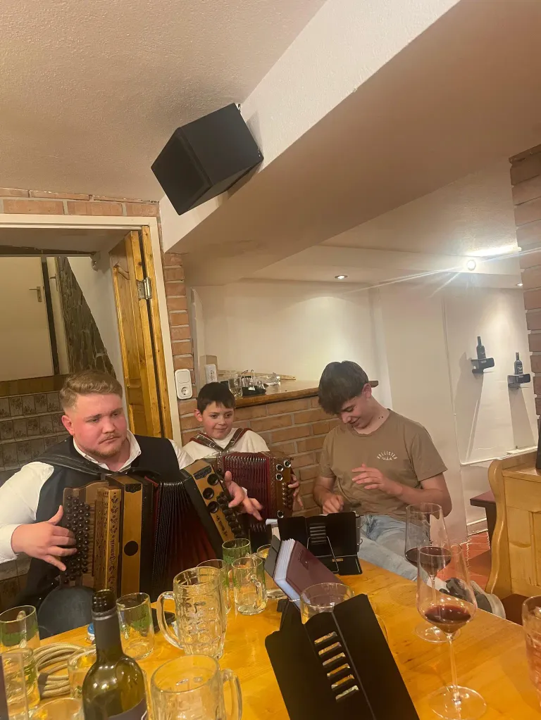 Three men sit at a table playing accordions. A man plays while another watches and smiles. They have wine glasses and a menu on the table. A speaker is mounted on the ceiling.