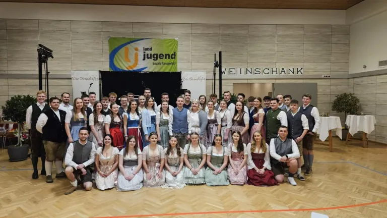 A group of people are posing for a photograph in a hall. They are wearing traditional clothing. A banner is hanging on the wall behind them.