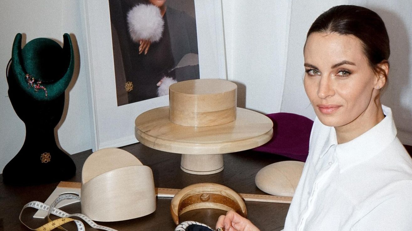 Eine hölzerne Hut-Vitrine mit mehreren Hüten und einem Maßband auf einem Tisch, eine Frau in der Nähe und ein Bilderrahmen im Hintergrund.