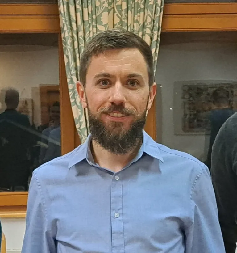 A man with a beard and mustache smiles in front of a window with curtains. He wears a light blue shirt. Behind him, another person's arm is visible.