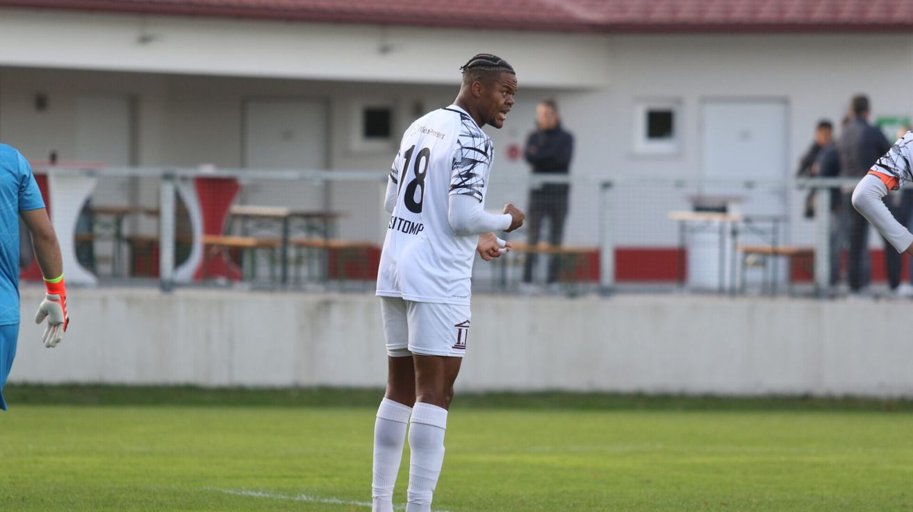 Ein Fußballspieler in einem weißen Trikot mit der Nummer 18 steht auf dem Feld und scheint zu feiern. Hinter ihm ist ein Gebäude mit einem roten Dach zu sehen, und eine Person steht in der Nähe eines Zauns.