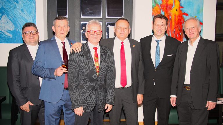 Four men in formal attire stand together, two wearing ties, one holding a drink. They seem to be attending an event, with a colorful painting in the background.