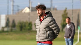 Ein Mann mit Bart steht auf einem Rasenfeld, trägt eine graue Daunenjacke mit Kapuze, ein rotes Unterhemd und blaue Jeans. Er schaut nach links, vielleicht beobachtet er etwas. Im Hintergrund ist eine verschwommene Ansicht eines Gebäudes und Bäume.