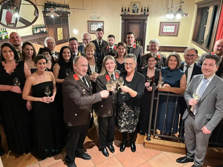 A group of people dressed in formal attire stand in a room, holding wine glasses, and appear to be celebrating an event. A woman in the center holds a glass of wine with both hands. Behind them, a few frames are mounted on the wall.