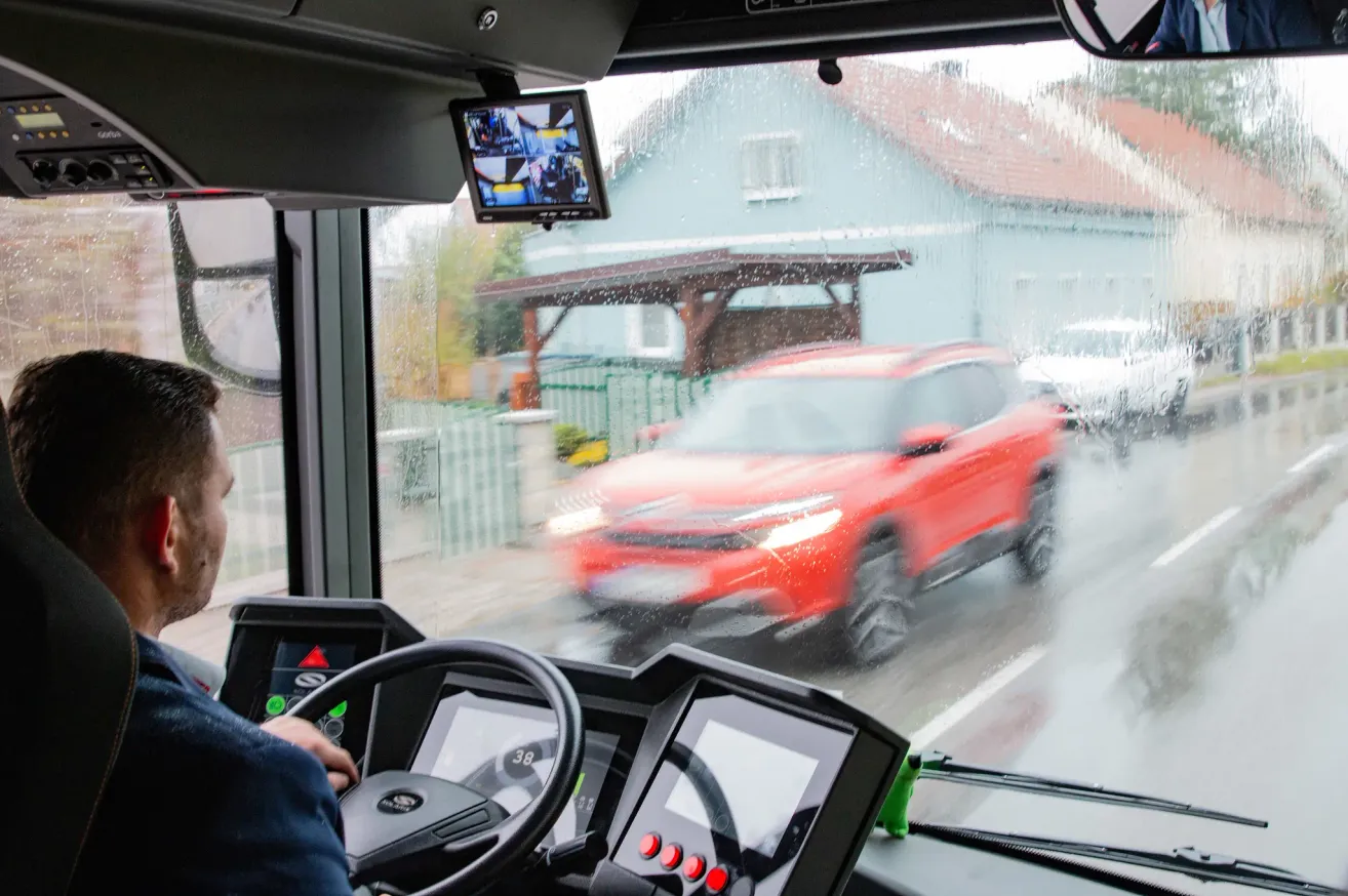 Von innen in einem Bus navigiert ein Fahrer mit einem roten SUV, der sich draußen in Bewegung befindet. Eine Rückfahrkamera ist über dem Armaturenbrett montiert.