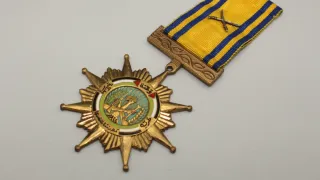 Eine goldene Sternmedaille mit einem gelben und blauen Band wird gezeigt. Der Stern hat einen grünen Kreis mit einem Soldaten und Weizengarben im Zentrum.