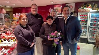 Fünf Personen stehen zusammen in einem Raum mit roter Wand und Blumen. Die Frau in der Mitte hält einen Blumenstrauß. Hinter ihnen ist ein Schild mit Text und Trophäen auf einem Regal.