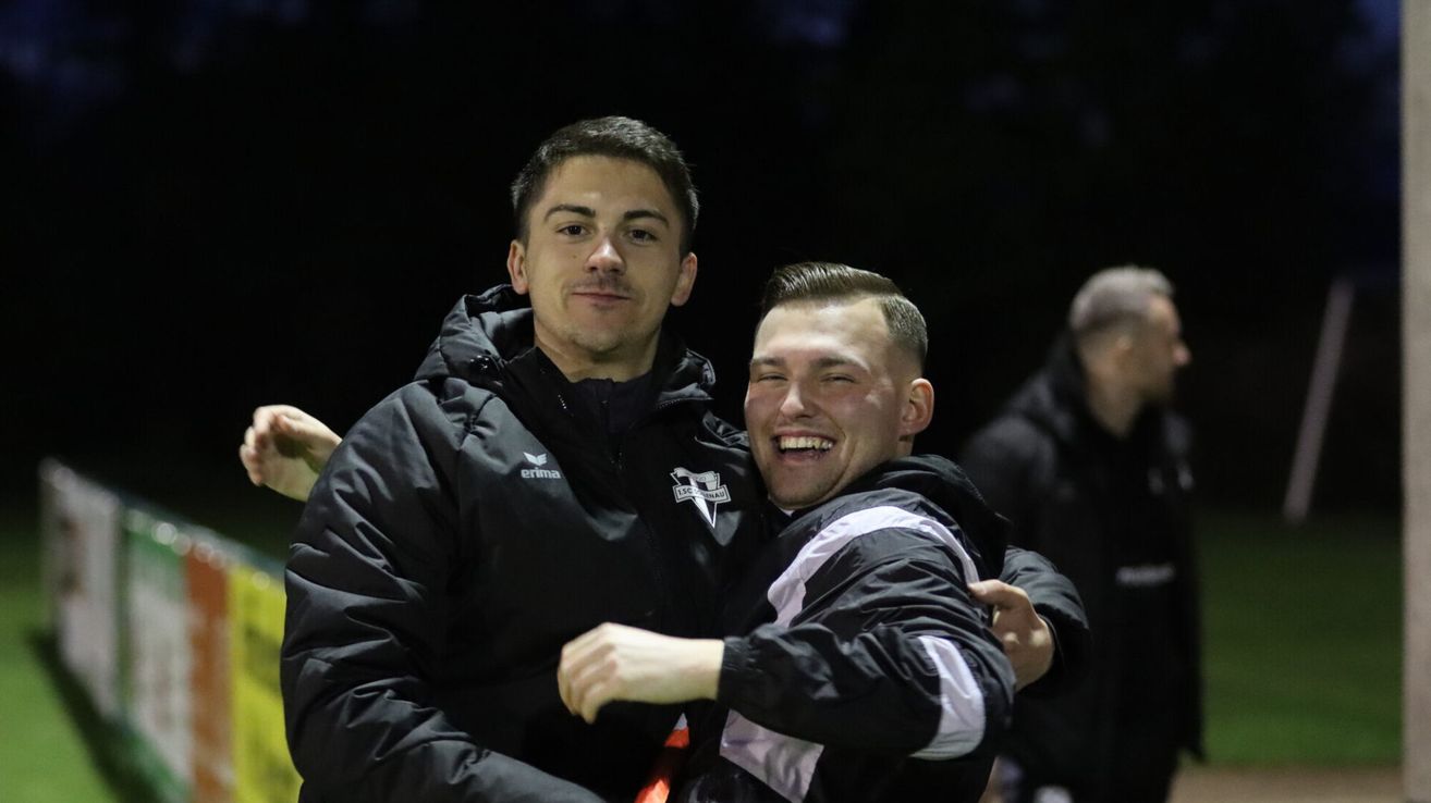 Zwei Männer in schwarzen Jacken umarmen sich lächelnd. Einer trägt ein 'erima'-Logo, und sie stehen in einem schwach beleuchteten Bereich.