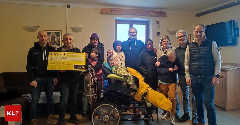 Eine Gruppe von Menschen posiert für ein Foto, zwei halten einen Scheck, einer im Rollstuhl, ein Baby in der Nähe und andere lächeln in die Kamera.