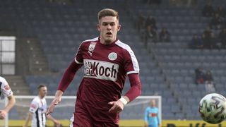 A soccer player wearing a maroon jersey with white accents runs on the field. The jersey features the words 'BAUKOCH' and a logo. In the background, there are empty seats and a blurred figure in blue.