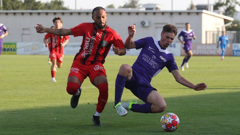Zwei Fußballspieler in roten und lila Trikots kämpfen auf dem Feld. Der Spieler in Rot jagt den Ball, während der Spieler in Lila verteidigt. Im Hintergrund laufen andere Spieler.