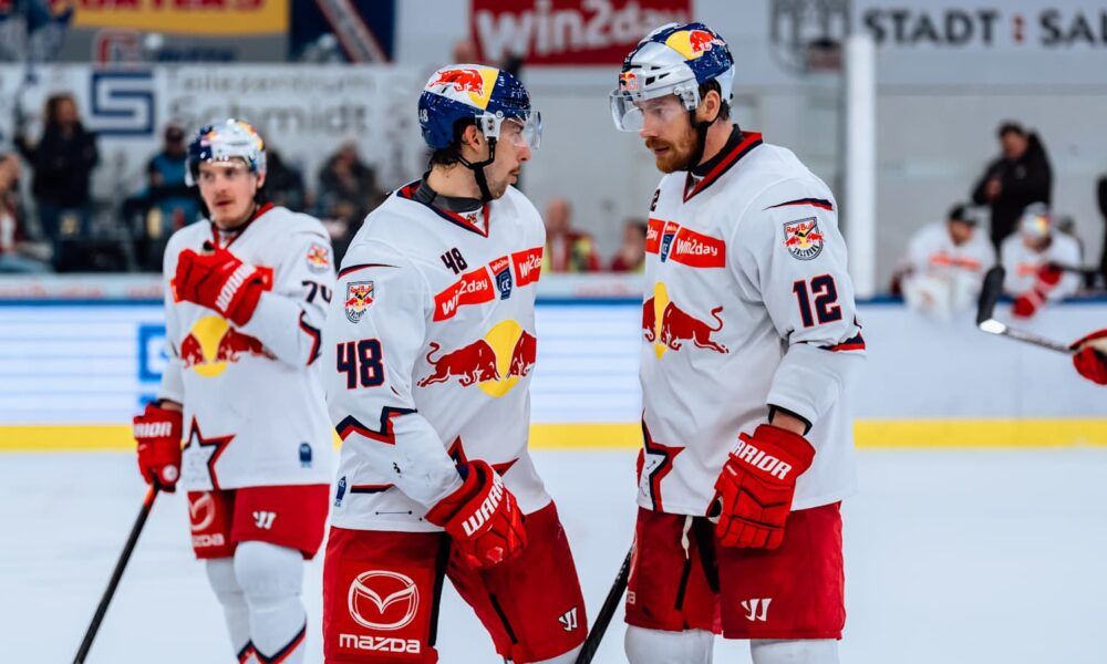 Three hockey players stand on the ice rink, wearing white jerseys with red accents. Two of them are conversing. Their jerseys feature the number 48, the Red Bull logo, and the word Warrior. The rink has a glass barrier and a sponsor's sign.