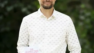 Ein Mann mit Bart steht in einem Garten und hält ein Buch mit dem Titel 'Die Kunst der Ausrede'. Er lächelt und trägt ein weißes Hemd mit kleinem Druck. Der Hintergrund ist unscharf mit viel Grün.