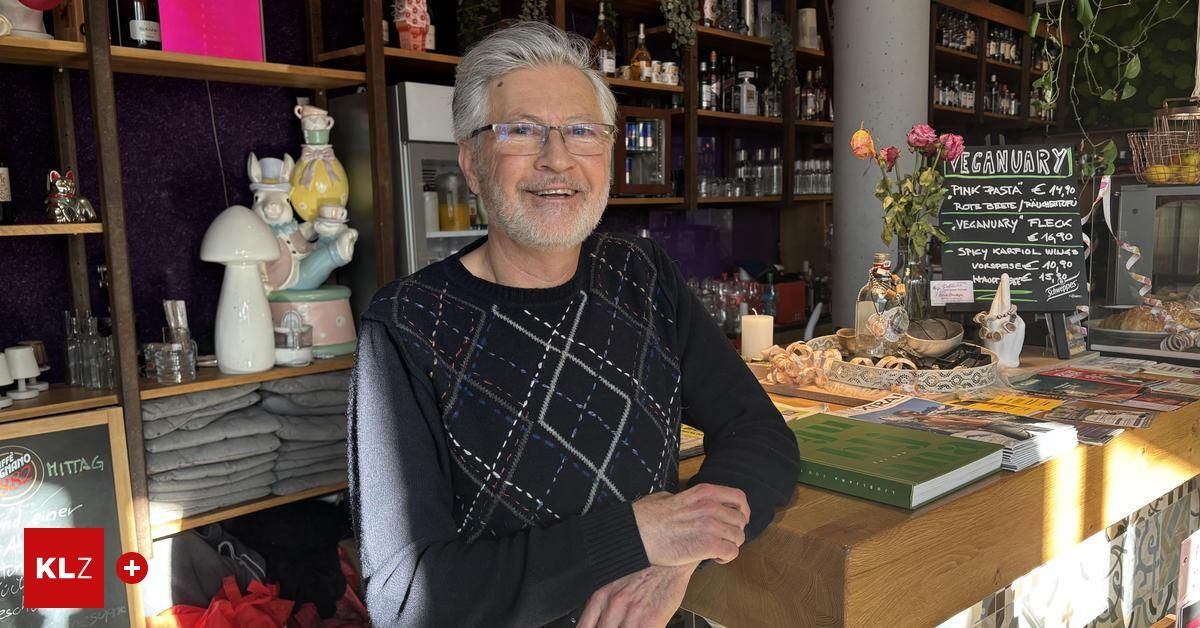 A man with glasses stands smiling in a shop, with shelves of bottles and jars behind him. In front, there is a table with a green book, flowers, and a bowl.