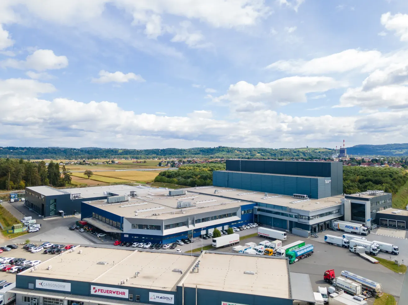 Luftaufnahme eines großen Industriebetriebs mit zahlreichen geparkten Lastwagen davor, umgeben von einer ländlichen Landschaft mit Feldern und Hügeln im Hintergrund.