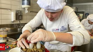 A chef in a kitchen wearing a white uniform, hat, and gloves, is preparing a cake while looking focused at the cake.