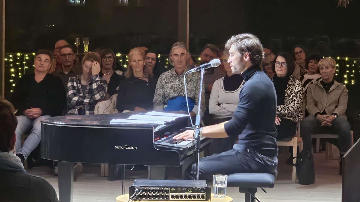 A pianist plays on a Dutch Grand piano with a microphone in front of an audience. Some people are looking at him while others are looking at something else.