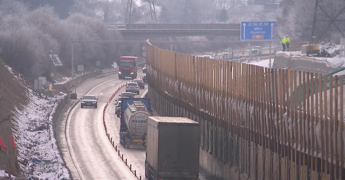 A highway with several cars and trucks driving on a snowy road. A tall wooden fence is on the right, and a bridge is in the distance.