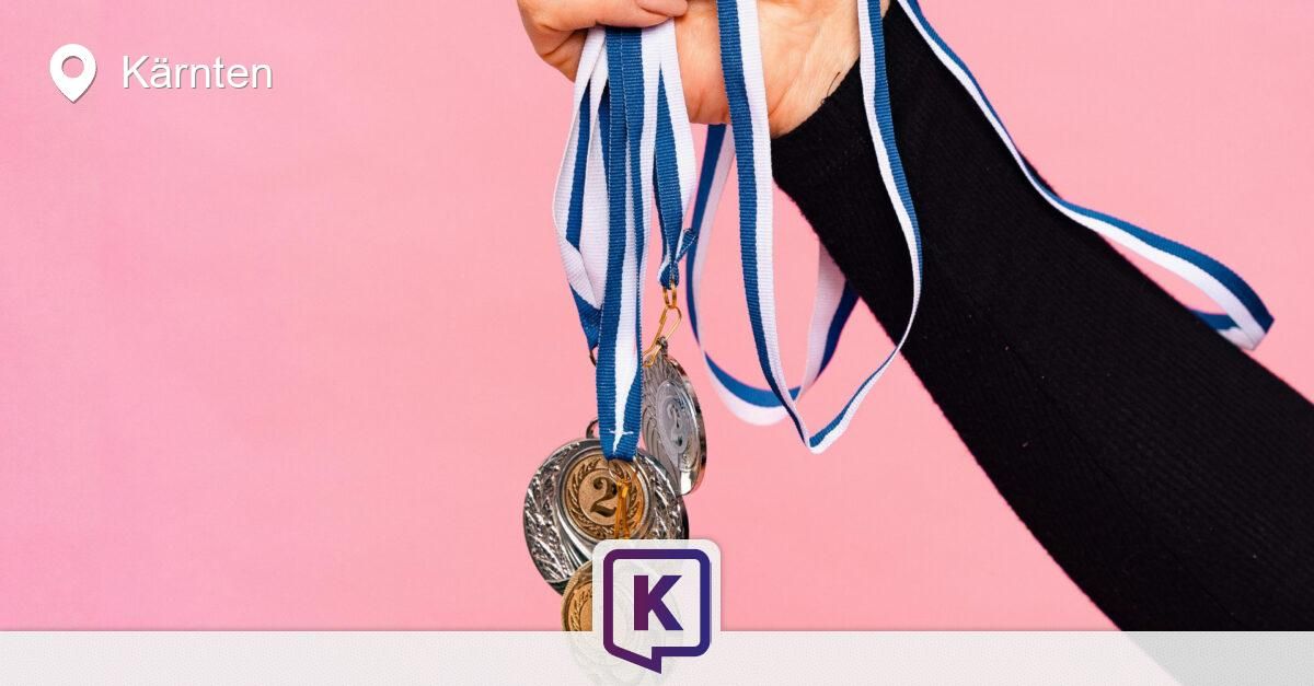 A hand holds three medals with blue and white ribbons against a pink background. The medals are gold, silver, and bronze.