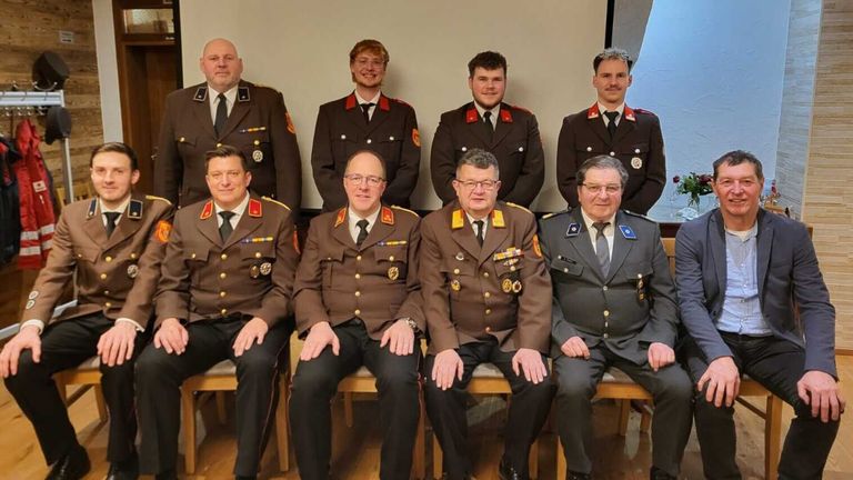 A group of men in military uniforms are seated on chairs, smiling and posing for a photo. Behind them, there is a white wall.