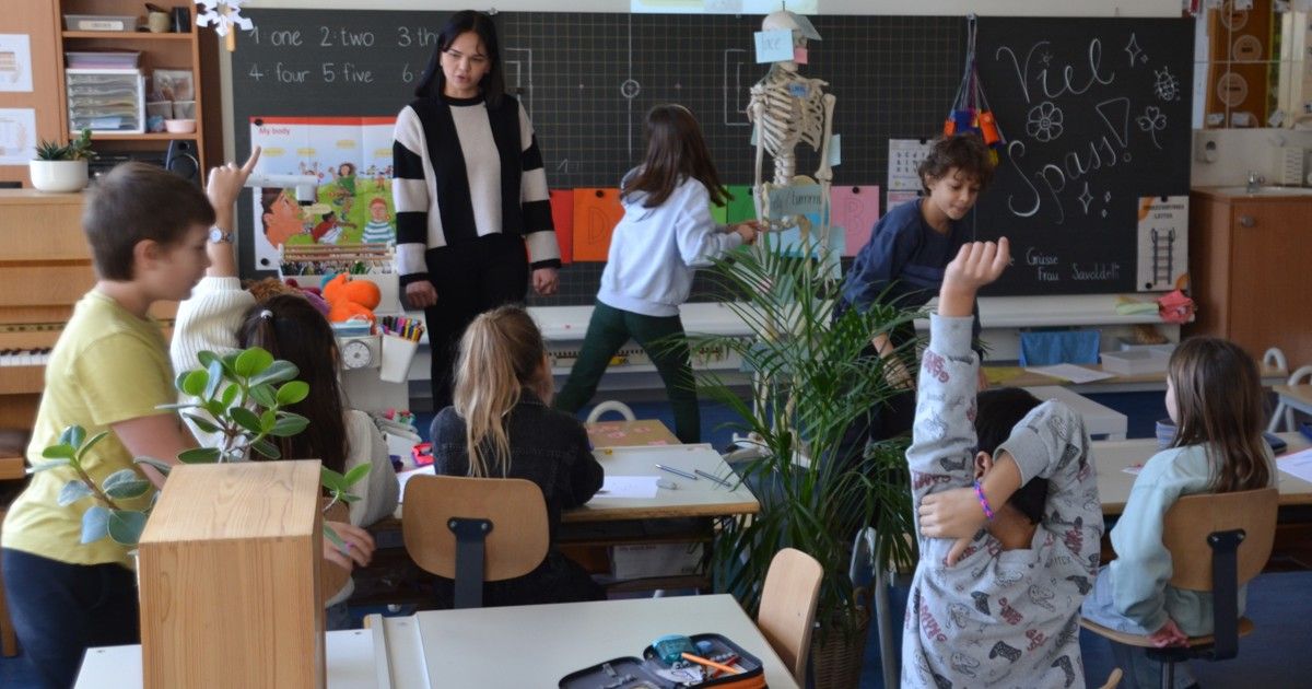 A woman in a black and white sweater is teaching a classroom of children. The children are standing, some with their hands raised, and there is a skeleton model and a blackboard in the background.