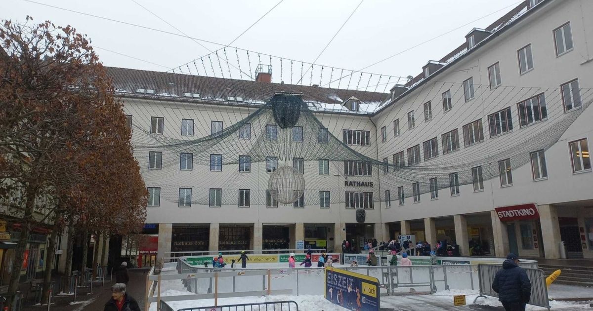 A building with a large canopy over an ice skating rink, children and adults ice skating. The building has many windows and is labeled Rathaus.