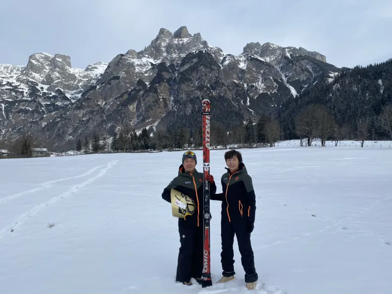 Zwei Personen stehen im Schnee, halten Skier und lächeln. Sie sind in Skiausrüstung gekleidet. Hinter ihnen befindet sich eine bergige Landschaft mit Schnee.