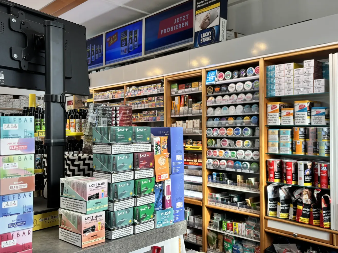 A store displays various products, including Lost Mary and Salt e-liquids. The store has wooden shelves, a counter, and a large monitor displaying a promotional advertisement.