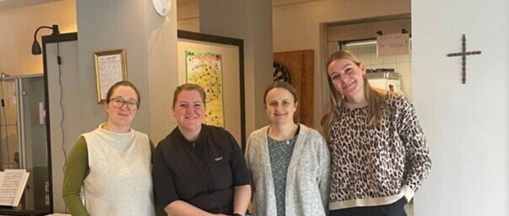 Four women stand close together in a room, smiling. Behind them, there are framed pictures and a white wall. A door with a sign is on the right.