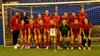 A women's soccer team is posing for a picture with a trophy on a field, holding a certificate that says 'Urkunde' and '1. Platz'. They are wearing red jerseys with sponsor logos.