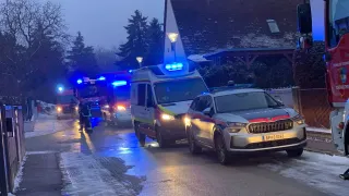 Emergency vehicles including ambulances and police cars are parked on a snowy street in front of a house. The scene is under blue lights.
