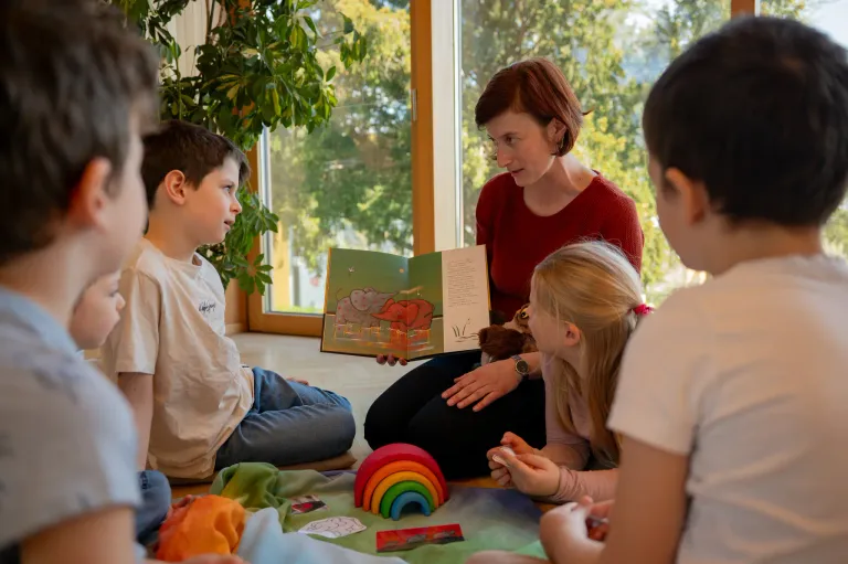 A woman reads a book to a group of children sitting in a circle, with toys and a rainbow nearby.