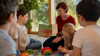A woman reads a book to a group of children sitting in a circle, with toys and a rainbow nearby.