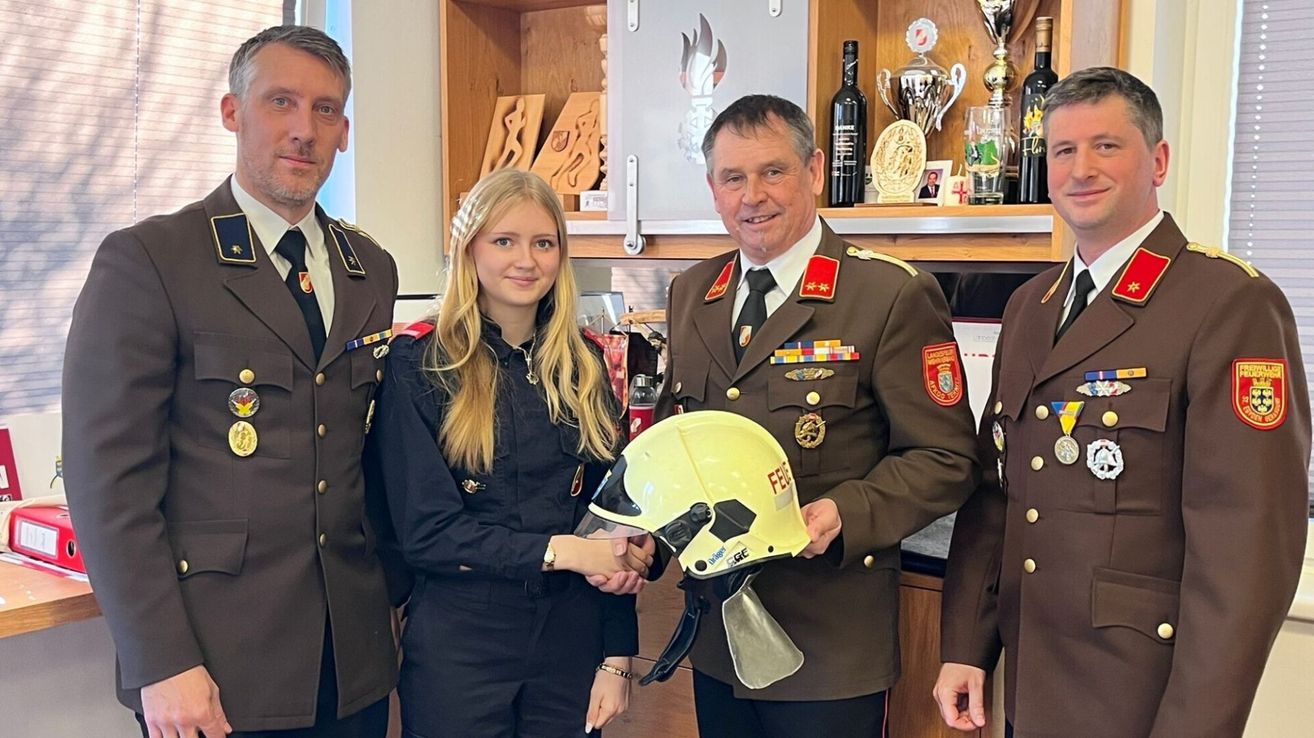 Three people stand in front of a wooden shelf. A woman and a man in uniforms, one holding a yellow helmet, and a man in a military uniform.