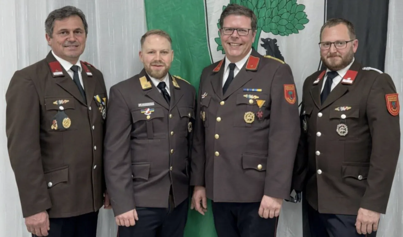 Four men in military uniforms are standing together in front of a green and white flag. They are all smiling.