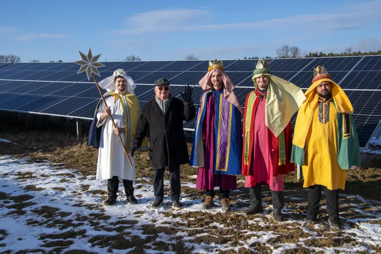 Five people dressed in colorful costumes stand in front of a solar panel installation. They appear to be posing for a photo, with two of them wearing crowns.