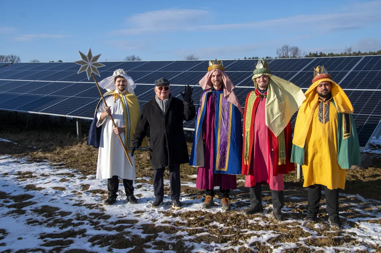 Five people dressed in colorful costumes stand in front of a solar panel installation. They appear to be posing for a photo, with two of them wearing crowns.