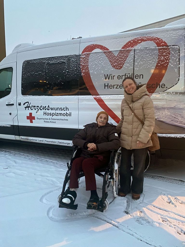 An elderly woman in a wheelchair stands outside a white van with red text reading Herzenswunsch Hospizmobil in snowy weather. She is smiling with another woman in a coat.