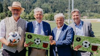 Four men pose in a field holding soccer balls and signs that say Danke Peter and Danke Daniel. The man on the left wears a hat and glasses. Behind them is a soccer net.