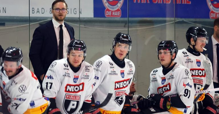 Three hockey players in NEON jerseys are sitting on the bench, looking at the camera. One wears number 17, and they all wear helmets. Behind them, a man in a suit looks at them.