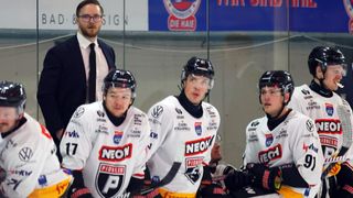 Three hockey players in NEON jerseys are sitting on the bench, looking at the camera. One wears number 17, and they all wear helmets. Behind them, a man in a suit looks at them.
